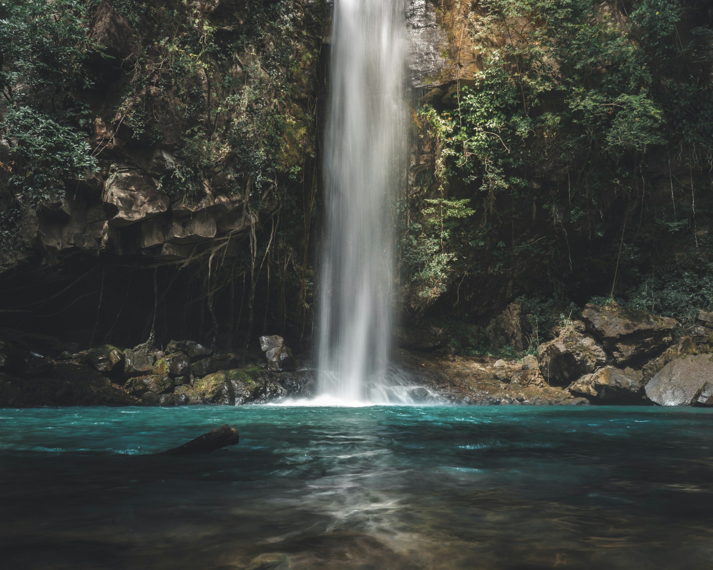 Tall waterfall cascading into a pool of water surrounded by rocks and lush green foliage.
